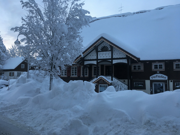 KI generiert: Das Bild zeigt ein Gebäude im Winter, stark eingeschneit, mit einem traditionellen Design und der Aufschrift "Hofladen". Die Umgebung ist ebenfalls mit tiefem Schnee bedeckt, und ein Baum im Vordergrund ist schwer von Schnee bedeckt.