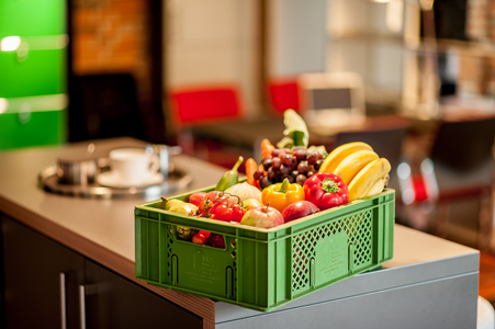 KI generiert: Das Bild zeigt eine grüne Kiste voller bunter Früchte und Gemüse, die auf einem Küchentresen steht. Im Hintergrund befindet sich eine gemütlich eingerichtete Wohnküche.