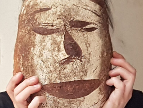 KI generiert: Das Bild zeigt eine Person, die eine große Brotlaib-Maske vor ihrem Gesicht hält. Das Brot ist so gestaltet, dass es Gesichtszüge aufweist.