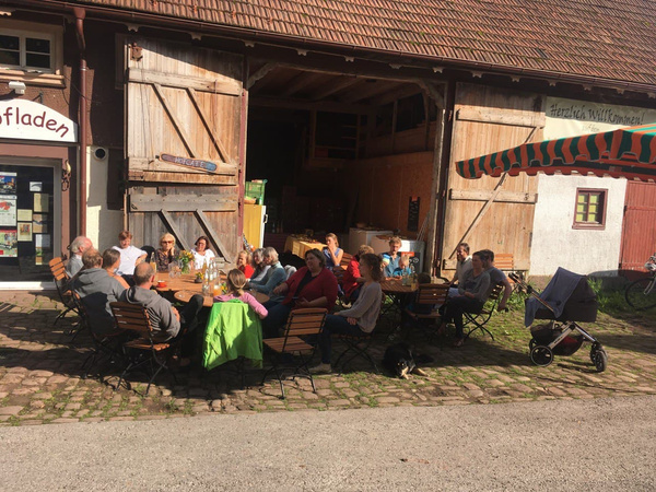 KI generiert: Eine Gruppe von Menschen sitzt draußen an Tischen vor einem Gebäude, das wie ein lokales Café oder Laden aussieht. Sie genießen offenbar das sonnige Wetter und die Gesellschaft.