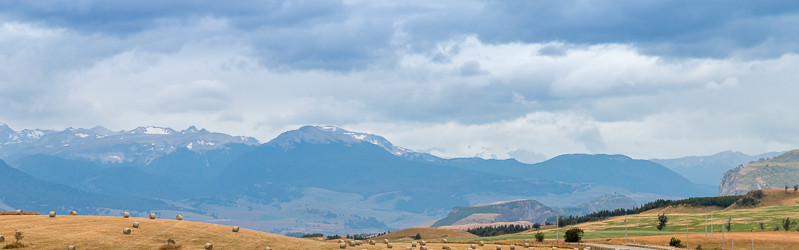 KI generiert: Das Bild zeigt eine lange, schlängelnde Straße in einer ländlichen Landschaft, gesäumt von Feldern mit Heuballen. Im Hintergrund erheben sich majestätische Berge unter einem bewölkten Himmel.