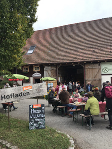 KI generiert: Das Bild zeigt einen belebten Bauernhofladen während eines Herbstfestes, wo Menschen an Bänken sitzen und feiern. Ein Willkommensschild für das Fest ist sichtbar, und der Laden wirkt einladend und geschäftig.