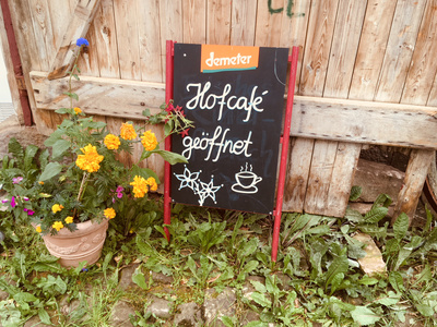 KI generiert: Das Bild zeigt ein Schild vor einem rustikalen Holzzaun, das auf ein geöffnetes Hofcafé hinweist. Neben dem Schild steht ein Blumentopf mit bunten Blumen.