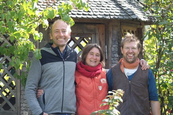 KI generiert: Das Bild zeigt drei lächelnde Personen, die Arm in Arm vor einem rustikalen Holzgebäude stehen, umgeben von grüner Vegetation. Sie wirken entspannt und freundlich in einer naturnahen Umgebung.