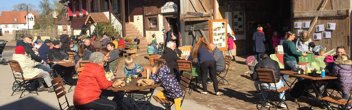 KI generiert: Mehrere Menschen sitzen bei sonnigem Wetter an Tischen vor einem Hofladen.
