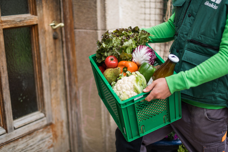 KI generiert: Ein Mensch hält eine grüne Kiste voller frischem Gemüse und Obst sowie eine Flasche Saft. Die Person steht vor einer Tür, möglicherweise bei einer Lieferung.