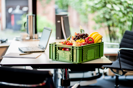 KI generiert: Das Bild zeigt einen Tisch in einem Büro, auf dem ein Laptop, eine Mappe und ein Korb mit verschiedenen Früchten und Gemüse stehen. Der Korb enthält unter anderem Bananen, Äpfel und Paprika.