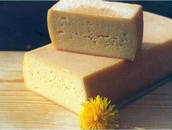 KI generiert: Ein Käselaib und ein Stück davon auf einem Holzbrett, daneben eine gelbe Blume.