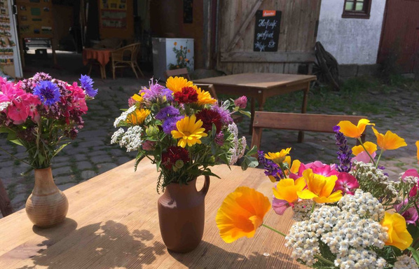 KI generiert: Das Bild zeigt drei Vasen mit bunten Blumensträußen auf einem Holztisch im Freien. Im Hintergrund sieht man einen Innenhof mit offenen Türen und Tischen.