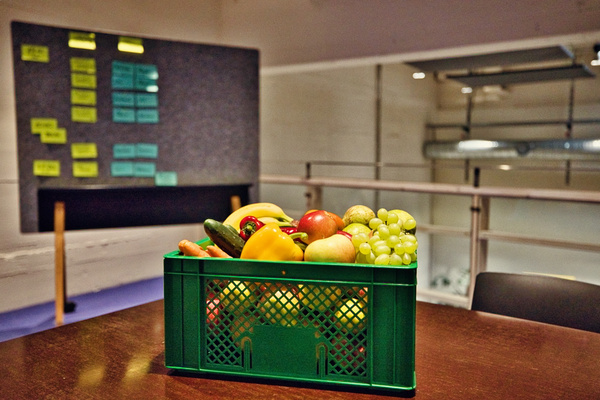 KI generiert: Das Bild zeigt eine grüne Kiste voller verschiedener Obst- und Gemüsesorten auf einem Tisch. Im Hintergrund ist eine Pinnwand mit Notizzetteln zu sehen.