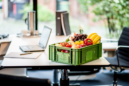 KI generiert: Das Bild zeigt einen modernen Büroarbeitsplatz mit einem Tisch, auf dem ein grüner Obstkorb mit verschiedenen Früchten und Gemüse steht. Im Hintergrund sind ein Laptop, eine Kaffeekanne und Büroutensilien sichtbar.