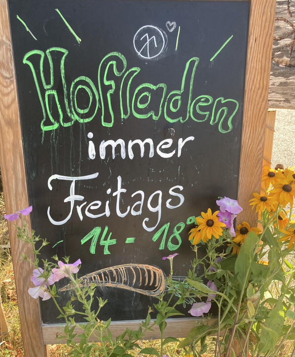 KI generiert: Tafel mit Öffnungszeiten: "Hofladen immer freitags 14 - 18 Uhr". Bunte Blumen umrahmen das Schild.