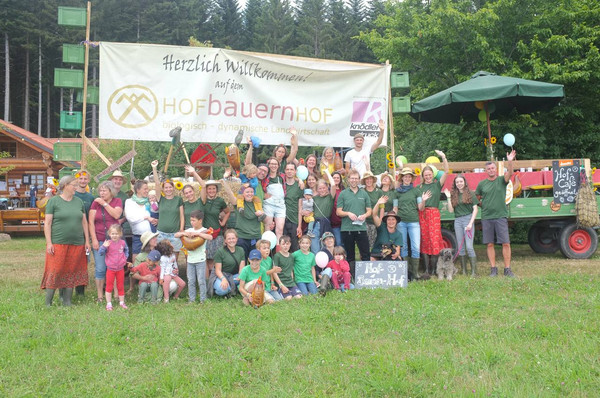 KI generiert: Das Bild zeigt eine Gruppe von Menschen, die vor einem Banner mit der Aufschrift "Hofbauernhof" in einer natürlichen Umgebung posieren. Die Szene vermittelt eine freundliche und einladende Atmosphäre eines Bauernhof-Events.