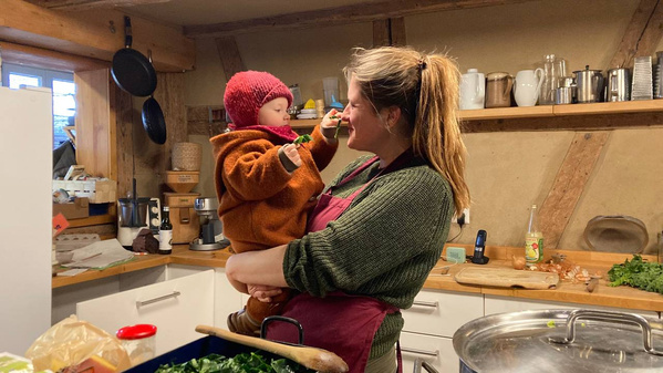 KI generiert: Das Bild zeigt eine Frau in einer Küche, die ein Kind auf dem Arm hält. Das Kind füttert die Frau mit einem Löffel.