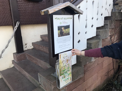 KI generiert: Das Bild zeigt ein Willkommensschild mit Informationen auf einem Hof, daneben hält eine Person einen Lageplan in der Hand. Das Schild hat einen Holzrahmen und darauf ist ein Bild von Kühen auf einer Weide abgebildet.