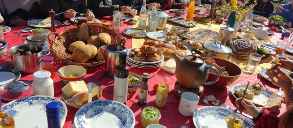 KI generiert: Das Bild zeigt einen reich gedeckten Tisch mit verschiedenen Speisen, darunter Brot, Aufstriche und Getränke. Mehrere Personen sitzen um den Tisch und genießen gemeinsam eine Mahlzeit.