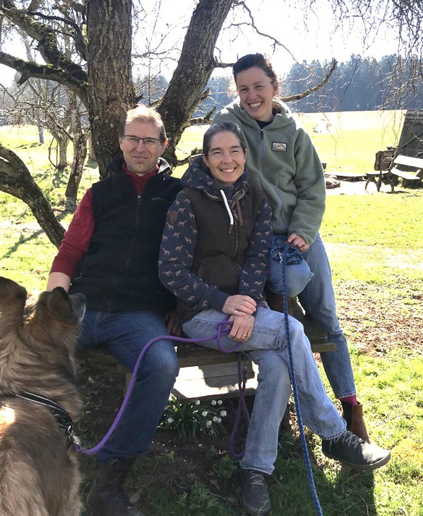 KI generiert: Drei Personen sitzen unter einem Baum auf einer Bank im Freien, mit einem Hund im Vordergrund.
