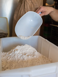 KI generiert: Das Bild zeigt eine Person, die mit einer Schaufel Mehl in einen Behälter umfüllt. Im Hintergrund ist eine braune Papiertüte zu erkennen.