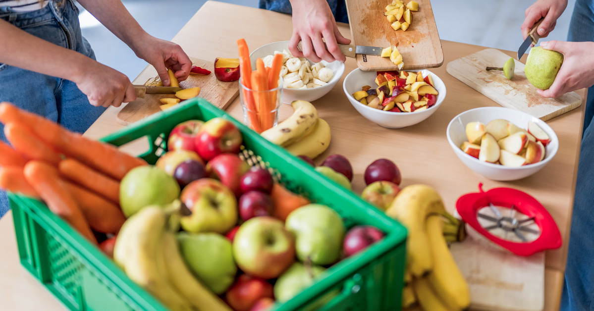 KI generiert: Menschen schneiden Obst neben einer Kiste voller Obst und Gemüse auf einem Tisch.