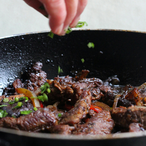 KI generiert: Wok mit gebratenem Fleisch und Gemüse; eine Hand gibt grüne Kräuter hinzu.