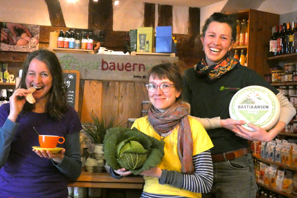 KI generiert: Drei Personen zeigen Lebensmittel wie Kohl und Käse in einem Laden. Text: "BASTIAANSEN jong".