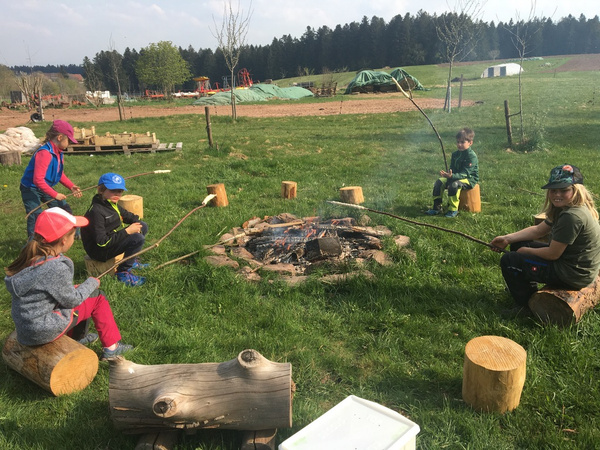 KI generiert: Das Bild zeigt eine Gruppe von Kindern, die um ein Lagerfeuer auf einer Wiese sitzen und Stöcke mit Teig über die Flammen halten. Im Hintergrund ist eine ländliche Landschaft mit Bäumen und landwirtschaftlichen Geräten zu sehen.