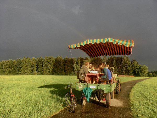 KI generiert: Auf dem Bild sitzt eine Gruppe von Menschen auf einem von einem Traktor gezogenen Wagen mit einem gestreiften Dach, inmitten einer ländlichen Landschaft, während im Hintergrund ein Regenbogen zu sehen ist. Das Bild vermittelt eine gemütliche und idyllische Atmosphäre.